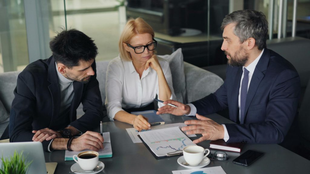 An image of 3 leaders looking at a dashboard of metrics. They are sitting at a table with coffee and papers discussing the report in front of them.