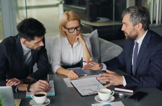 An image of 3 leaders looking at a dashboard of metrics. They are sitting at a table with coffee and papers discussing the report in front of them.